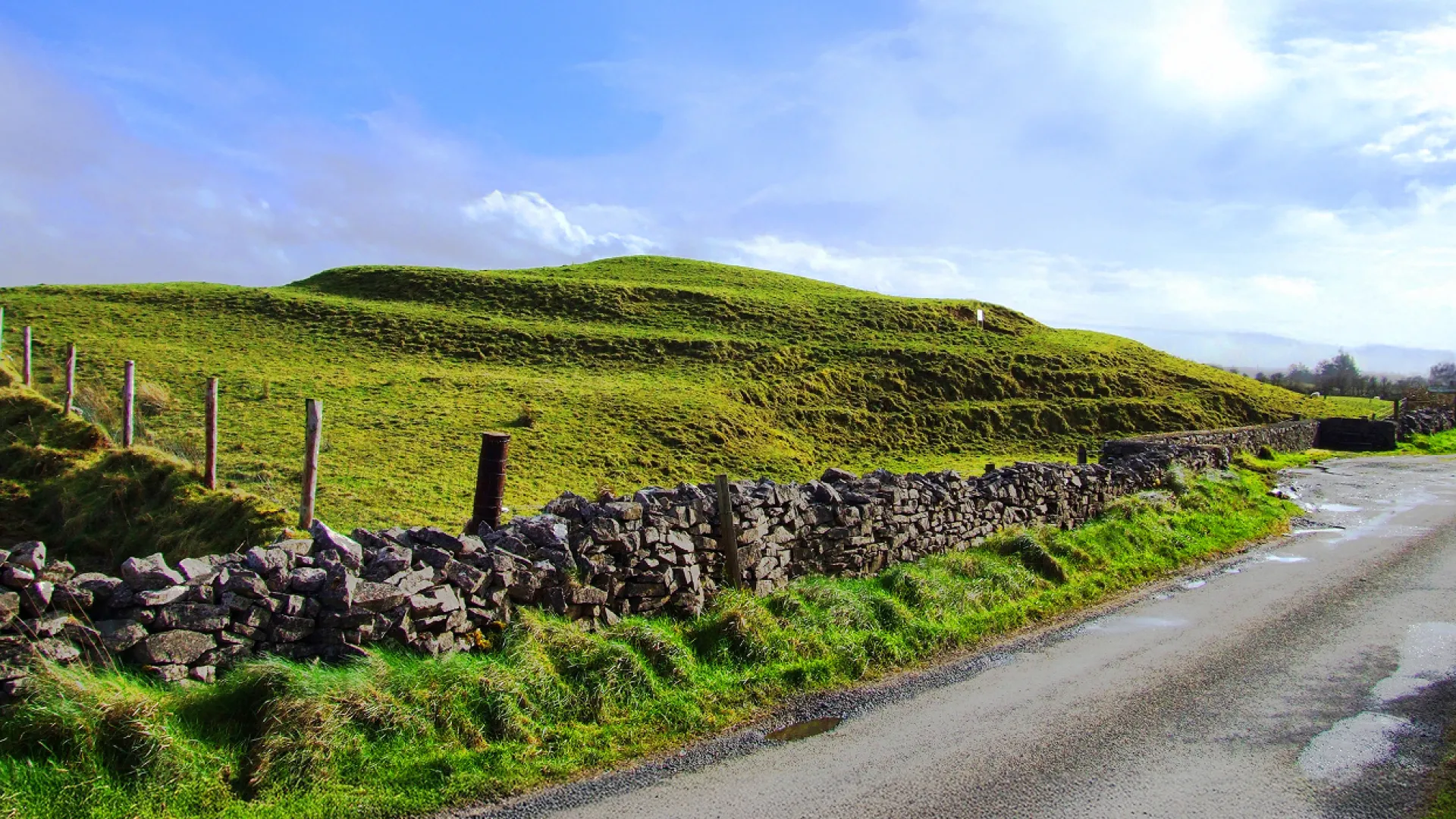 Verdant, undulating hills covered in lush grass, with a winding dirt road and stone walls running along the landscape.